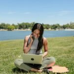 Smiling woman sits on grass by a lake, using a laptop and talking on the phone, enjoying the outdoors.