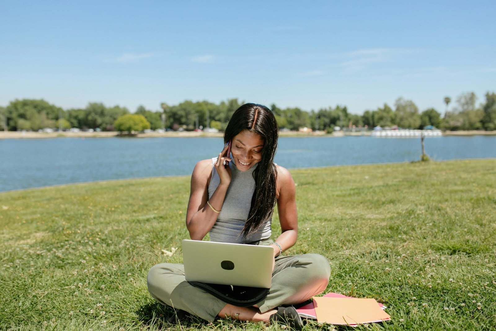 Smiling woman sits on grass by a lake, using a laptop and talking on the phone, enjoying the outdoors.